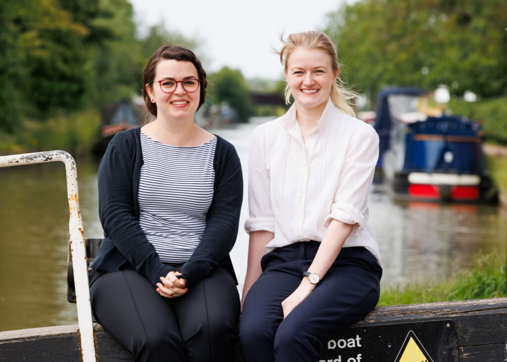 Image description: Lodders trainee solicitors Olivia Beeton and Lizzie Curnock. Olivia and Lizzie are sitting on a lock next to a canal, with a narrow boat out of focus behind them.